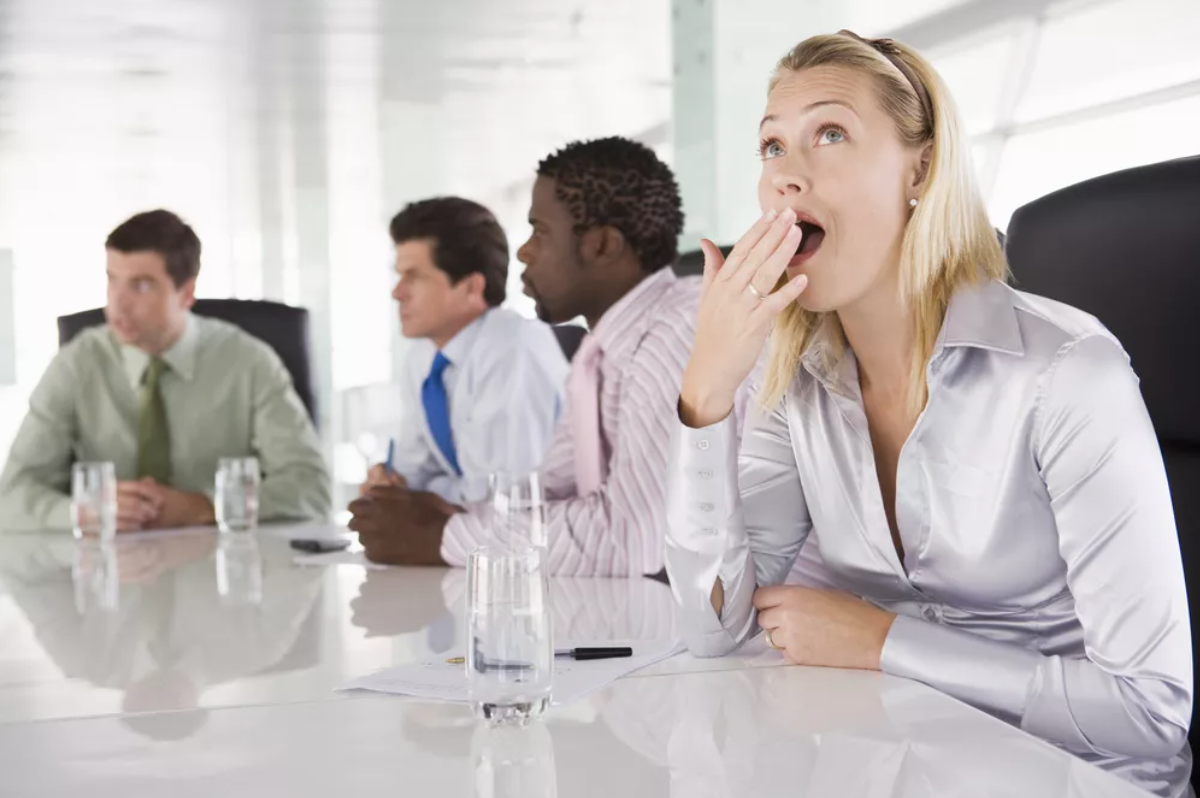 A photograph of a boardroom meeting. A white woman with blonde hair yawns in the foreground.